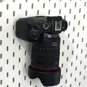 May include: A black Canon mirrorless camera with a 24-105mm lens attached, featuring a prominent red ring around the lens barrel and a black lens hood. The camera body displays various control dials, a hot shoe, and the Canon brand name on the lens.