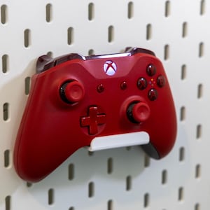 May include: A matte red wireless video game controller featuring a central Xbox logo button, two analog sticks, a directional pad, and four action buttons, mounted on a white wall-mounted storage pegboard panel with vertical slots.