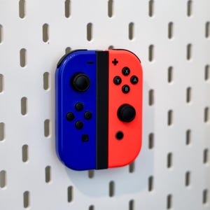 May include: A blue and red video game controller attached to a white pegboard. The left side is blue with a black joystick and directional buttons, while the right side is red with a black joystick and four action buttons labeled A, B, X, and Y.