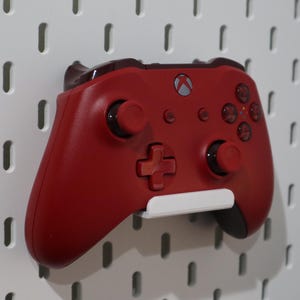 May include: A red Xbox controller mounted on a white pegboard. The controller has black buttons and a white bracket. The pegboard has evenly spaced, vertical slots.