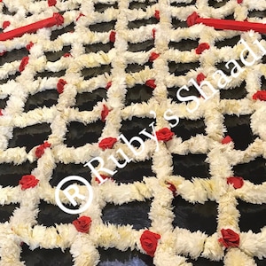 Puede incluir: Una guirnalda floral blanca con rosas rojas dispuestas en forma de cuadrícula sobre una superficie negra. El texto "Ruby's Shaadi" es visible en la imagen.