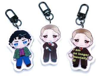 May include: Three cartoon character keychains with black metal key rings. The keychain on the left features a cartoon man with black hair wearing a green jacket, blue jeans, and a red and white plaid shirt. The keychain in the middle features a cartoon man with blonde hair wearing a brown and black plaid suit and holding a knife. The keychain on the right features a cartoon man with blonde hair wearing a brown and black plaid suit. The keychain on the right has the text "i am no longer mentally ill" printed on it.