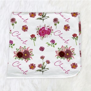Puede incluir: Manta de beb&eacute; blanca con un patr&oacute;n floral en tonos rosa y burdeos. La manta tiene el nombre "Charli" repetido en todo el dise&ntilde;o.