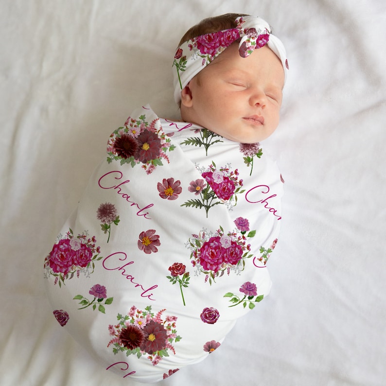 Puede incluir: Una manta de arrullo blanca con un patr&oacute;n floral en tonos rosa y burdeos. La manta tiene el nombre "Charli" impreso varias veces. Un beb&eacute; est&aacute; envuelto en la manta y lleva una diadema rosa con un lazo.