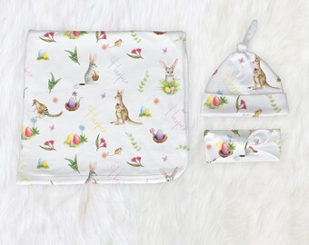 May include: A white baby blanket, hat, and headband set with a repeating pattern of colorful Easter eggs, kangaroos, and other Australian animals. The set is personalized with the name "Harper" in a variety of colors.