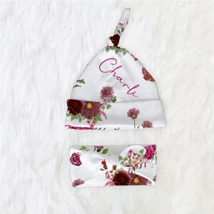 Puede incluir: Un gorro de beb&eacute; blanco con estampado floral y el nombre "Charli" bordado en rosa. El gorro est&aacute; atado en la parte superior con un nudo. Tambi&eacute;n se incluye una diadema a juego con estampado floral.