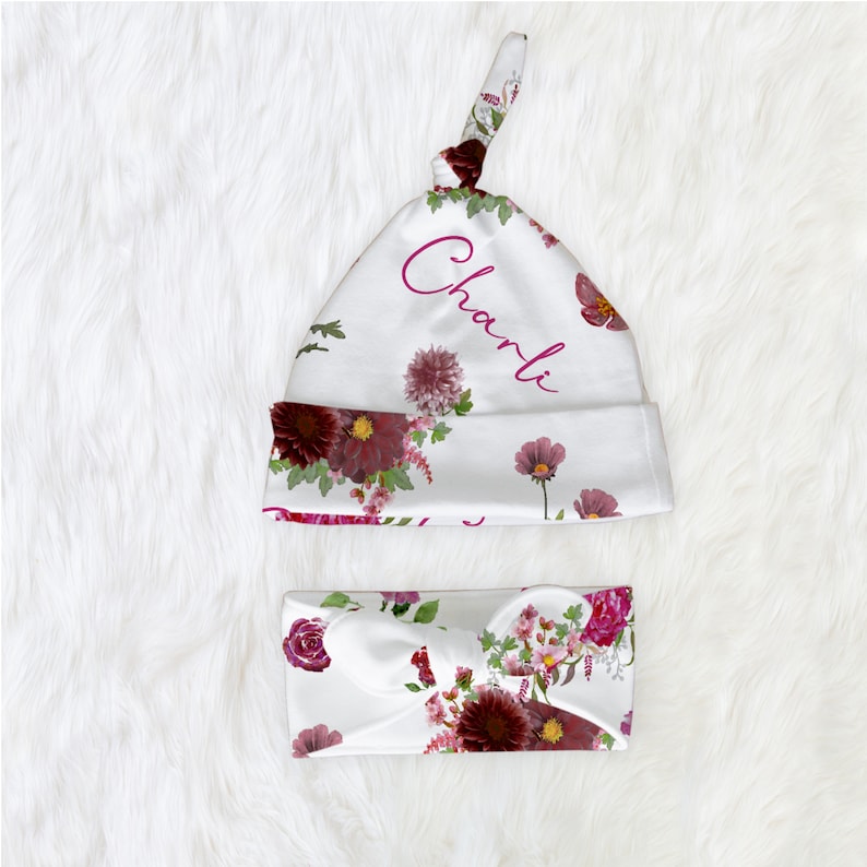 Puede incluir: Un gorro de beb&eacute; blanco con estampado floral y el nombre "Charli" bordado en rosa. El gorro est&aacute; atado en la parte superior con un nudo. Tambi&eacute;n se incluye una diadema a juego con estampado floral.