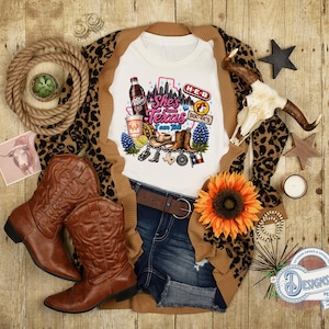 May include: A white t-shirt featuring a graphic print with the text 'She's from Texas I can Tell', a Dr Pepper bottle, a Buc-ee's logo, an H-E-B logo, a cowboy hat, boots, bluebonnets, and a Texas state outline. The shirt is layered with a leopard print cardigan.