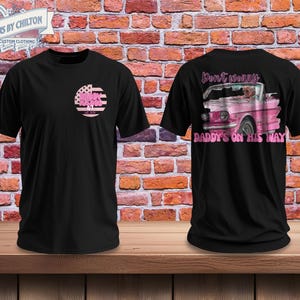May include: Black t-shirts with a graphic design. One shirt features a pink and white circular design with text, and the other displays a pink vintage car with the text "Don't worry, Daddy's on his way."