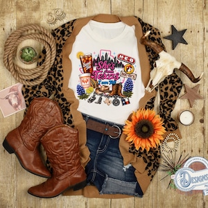 Può includere: Una t-shirt bianca con una stampa grafica con il testo She's from Texas I can tell, circondata da illustrazioni di un bicchiere di tè, un logo Buc-ee's, un cartello H-E-B, stivali da cowboy, un cappello, lupini e il profilo del Texas.