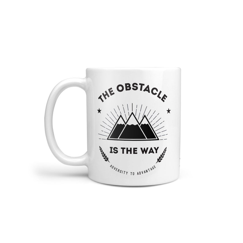 May include: White ceramic mug with a black graphic design featuring three mountains with a sunburst behind them. The text "The Obstacle Is The Way" is printed below the mountains. The text "Adversity To Advantage" is printed below the main text.