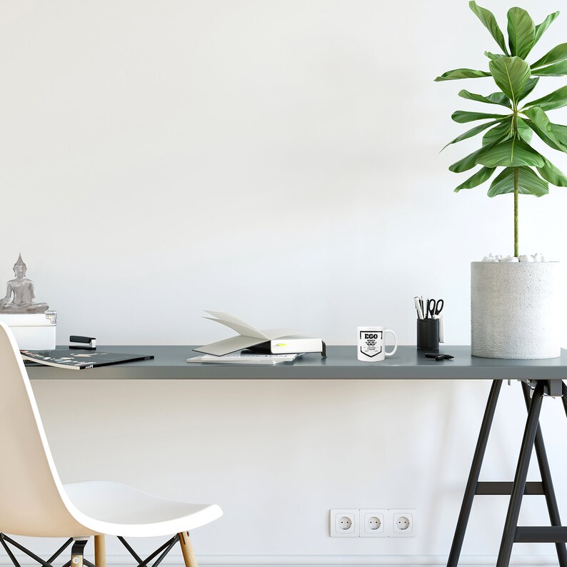 May include: White ceramic mug with black text that reads "Ego is the enemy". The mug is on a gray desk with a plant, a book, and a stapler.