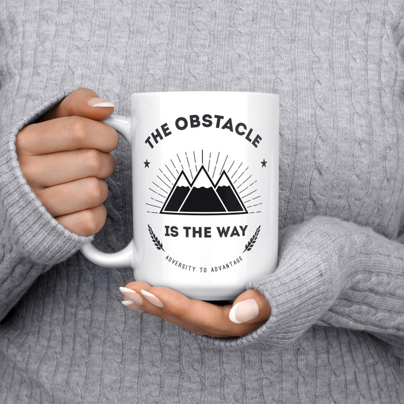 May include: White ceramic mug with a black graphic of three mountains with a sunburst behind them. The text "The Obstacle Is The Way" is printed below the mountains. The text "Adversity To Advantage" is printed below the text.