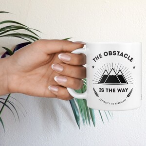 May include: White ceramic mug with a black graphic design featuring three mountains with a sunburst above them and the text "The Obstacle Is The Way" with the words "Adversity to Advantage" below the mountains.