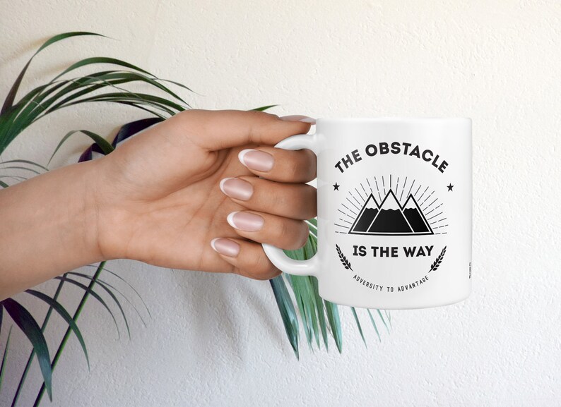 May include: White ceramic mug with a black graphic design featuring three mountains with a sunburst above them and the text "The Obstacle Is The Way" with the words "Adversity to Advantage" below the mountains.