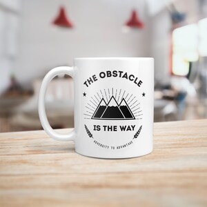 May include: White ceramic mug with a black graphic design featuring a mountain range with the text "The Obstacle Is The Way" and "Adversity To Advantage".