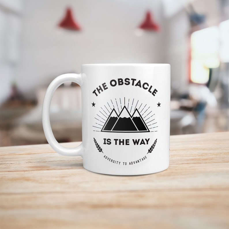 May include: White ceramic mug with a black graphic design featuring a mountain range with the text "The Obstacle Is The Way" and "Adversity To Advantage".