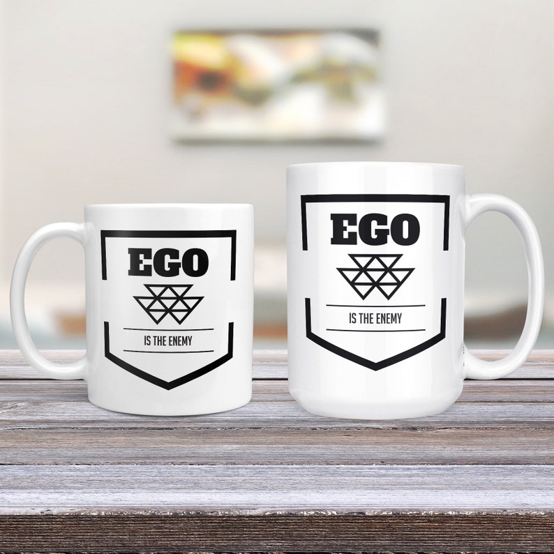 May include: Two white ceramic mugs with black text and a geometric design. The larger mug reads "EGO IS THE ENEMY" and the smaller mug reads "EGO IS THE ENEMY".