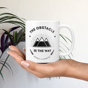 May include: White ceramic mug with a black graphic of three mountains with a sunburst behind them. The text "The Obstacle Is The Way" is printed below the mountains. The text "Adversity To Advantage" is printed below the main text.