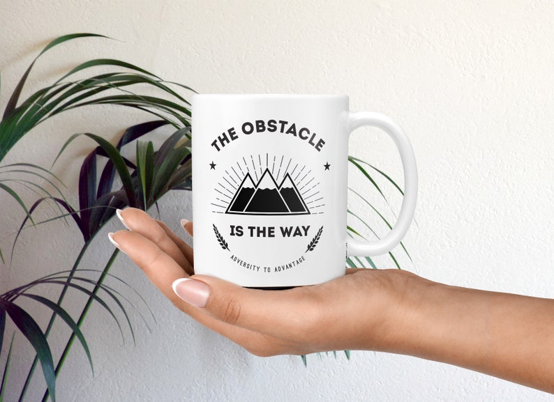 May include: White ceramic mug with a black graphic of three mountains with a sunburst behind them. The text "The Obstacle Is The Way" is printed below the mountains. The text "Adversity To Advantage" is printed below the main text.