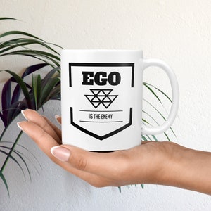 May include: White ceramic mug with a black graphic design. The graphic features a shield with the text "EGO IS THE ENEMY" inside.