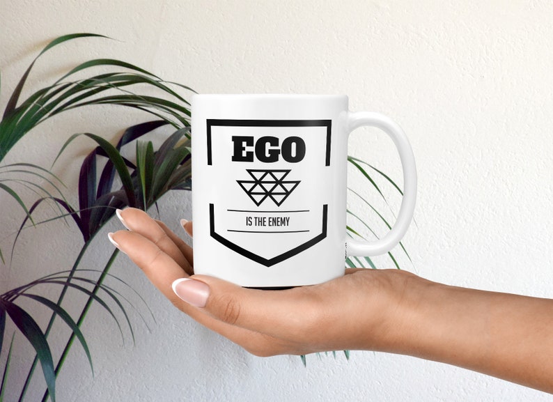 May include: White ceramic mug with a black graphic design. The graphic features a shield with the text "EGO IS THE ENEMY" inside.