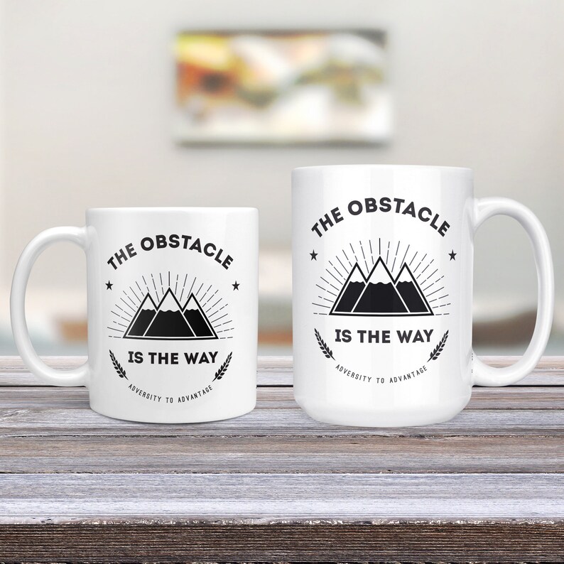 May include: Two white ceramic mugs with black text and graphics. The text on both mugs reads "The Obstacle is the Way" with the words "Adversity to Advantage" underneath. The graphic on the mugs is a stylized mountain range with a sunburst behind it.