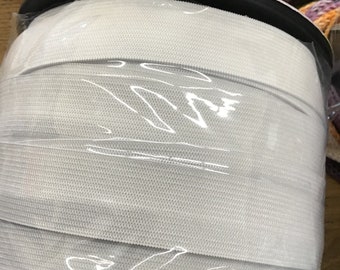 Puede incluir: Un rollo de cinta elástica blanca, bien enrollada y envuelta en plástico transparente. La cinta elástica parece tener aproximadamente 2,5 cm de ancho. El rollo está sobre una base de plástico negro. Se aprecia la textura de la cinta.