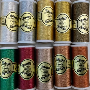 May include: A selection of ten spools of metallic thread in shades of silver, gold, bronze, and green. Each spool features a gold label with a butterfly design and text. The threads are tightly wound on the spools.