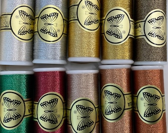 Puede incluir: Una colección de diez carretes de hilo metálico en varios colores, incluyendo plata, oro, bronce y verde. Cada carrete tiene una etiqueta dorada con un diseño de mariposa y texto. Los hilos están bien enrollados.