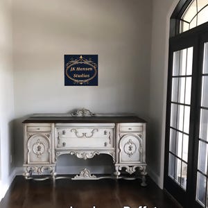 May include: A white and brown antique buffet with ornate carvings and a dark wood top. The buffet has drawers and cabinet doors. The image also includes a sign that says "JK Hansen Studios" and the text "Jacobean Buffets".