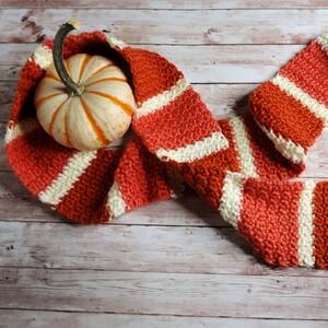 May include: A knitted scarf with orange and white stripes, folded and draped over a small white pumpkin.