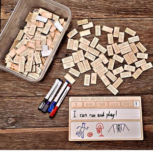 Puede incluir: Un kit de construcción de oraciones con palabras de vocabulario de madera y rotuladores sobre una superficie de madera. El kit incluye un recipiente de plástico transparente lleno de fichas, rotuladores y una pizarra con el texto "I can run and play!" e ilustraciones.