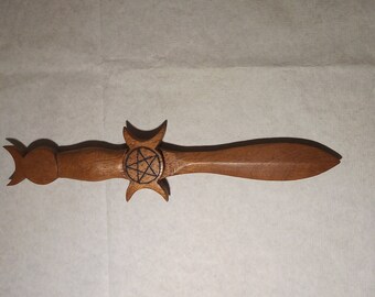 May include: A wooden dagger with a triple moon design and a pentagram in the center. The dagger is brown and has a sharp point.