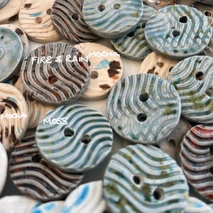 May include: A selection of ceramic buttons featuring different patterns and colours, including blue, white, brown, and green. The buttons have two holes and are labelled with names such as "Fire & Rain", "Mocha", "Moss", and "Delft".