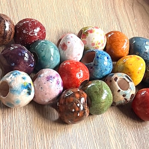 May include: A collection of round, glazed ceramic beads in various colours including red, blue, green, yellow, brown, purple, and pink. Each bead features a smooth, glossy finish with unique speckled or mottled patterns and a single central hole.