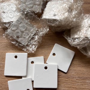 May include: White square ceramic tiles with a hole in the top corner, some wrapped in bubble wrap. The tiles are arranged on a light brown wooden surface. The tiles are a solid white colour.