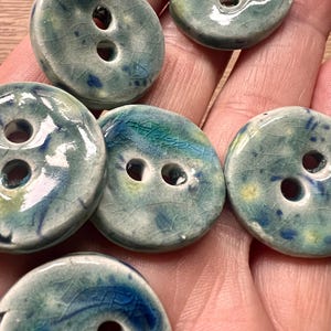 May include: A selection of handmade ceramic buttons in blue, green, and white hues. Each button features two holes and a crackle glaze. The round buttons vary slightly in size and colour, showcasing unique patterns.