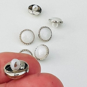 May include: Five white buttons with silver metal rims. The buttons have a raised, textured surface and a small hole in the centre.
