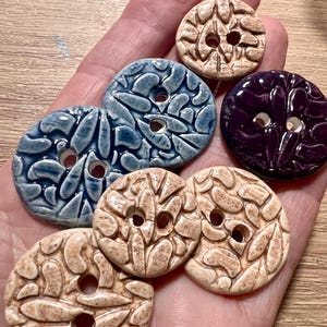 May include: A selection of handmade ceramic buttons in various colours. The buttons are round with two holes and feature a textured, organic design. Colours include shades of blue, beige, and purple. These buttons are ideal for sewing and craft projects.