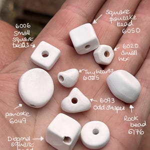 May include: A collection of white ceramic beads in various shapes and sizes, including square, round, pancake, and heart shapes. The beads are labelled with their corresponding product numbers and descriptions.