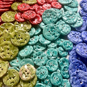 May include: A selection of handmade ceramic buttons in a range of colours. The round buttons have two or four holes and star or leaf patterns. Colours include red, green, teal, and blue. These decorative buttons are suitable for sewing and craft projects.