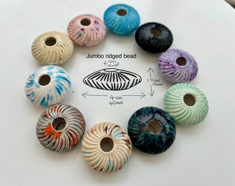 May include: A collection of 10 ceramic beads with a ridged design. The beads are 4 cm in diameter and have a 1.9 cm hole. The beads are in various colours, including blue, green, pink, purple, and black.