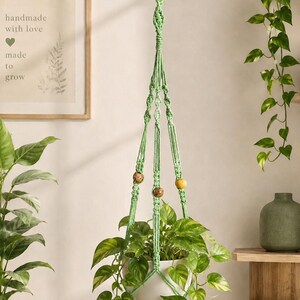 May include: A green macramé plant hanger with a wooden ring and decorative beads, holding a white ceramic pot filled with a leafy green plant. The image includes a framed print with the text "handmade with love made to grow."