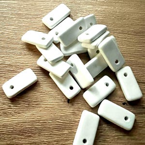 May include: A scattering of small, rectangular, white ceramic beads, each with a single hole. The beads are arranged on a wooden surface, highlighting their smooth texture and simple design. Suitable for jewellery making or craft projects.