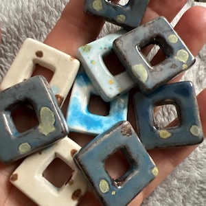 May include: A collection of eight ceramic square tiles with a hole in the centre. The tiles are decorated with a blue, white and brown glaze and have a speckled appearance.