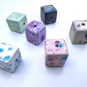 May include: Six ceramic cubes with different patterns and colours. The cubes are all about 5 cm in size and have a hole in the centre. The colours include light green, purple, pink, white with blue dots, grey, and beige with purple and brown dots.