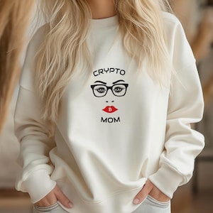 May include: Cream-colored sweatshirt with a graphic design featuring a face with glasses, red lips, and a Bitcoin symbol. The text "CRYPTO MOM" is above and below the face. The sweatshirt has long sleeves and a crew neck.