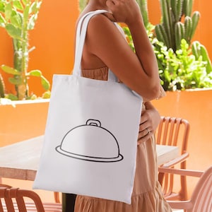 May include: A white tote bag featuring a black line drawing of a covered serving dish. The bag is held in front of a person. The background includes a wooden table, peach-colored chairs, and a bright orange wall with green plants.