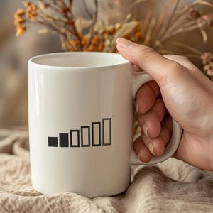May include: A white ceramic mug with a black signal strength graphic. The mug is held by a person's hand, with dried flowers in the background. The mug is a standard size for coffee or tea.